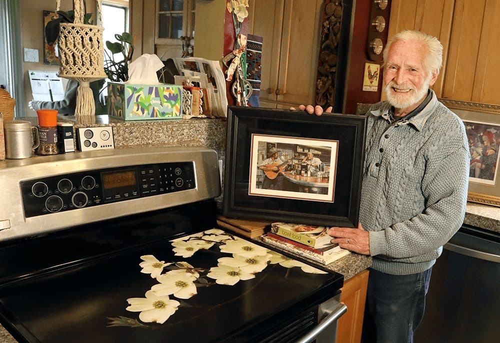 Artist Steven Saylor holding a framed painting beside a stove printed with dogwood flowers