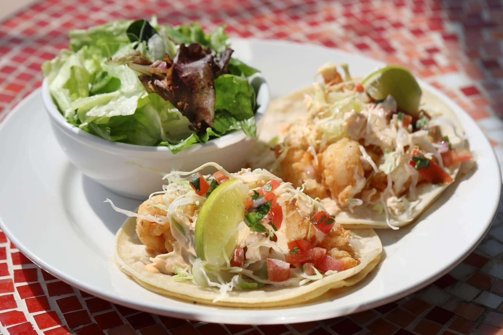 Crispy shrimp tacos with lime and side salad on a white plate, red mosaic table