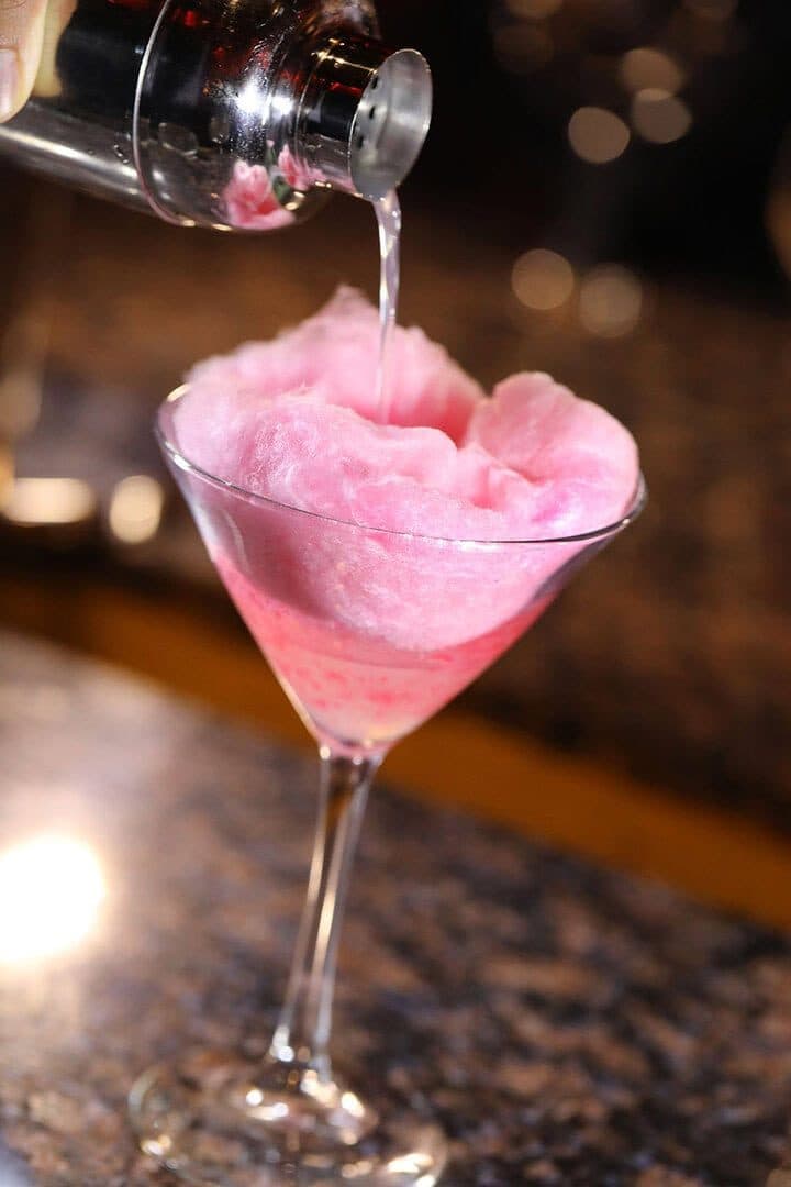 Cotton candy martini with liquid being poured over pink floss, dark bokeh bar