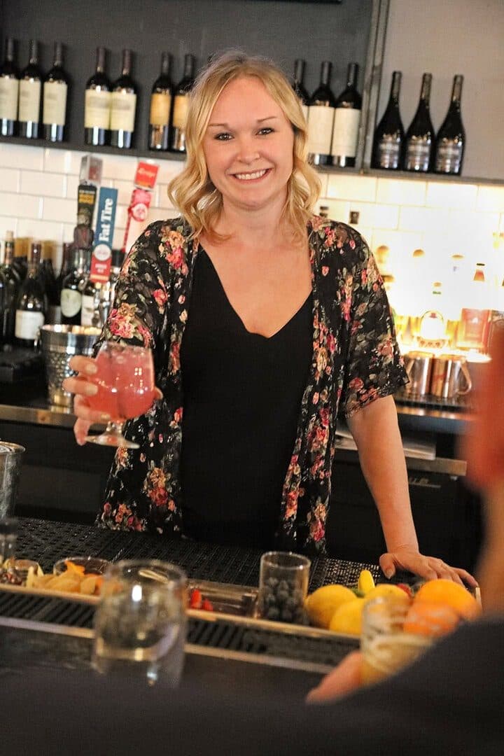 Woman at a bar raising a pink cocktail, wine bottles behind