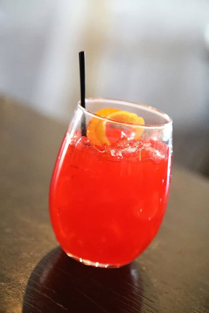 Red cocktail with orange garnish and straw in a stemless glass, dark background