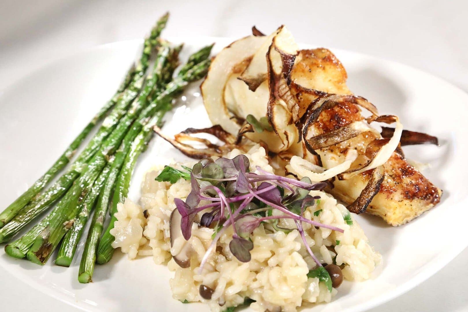Mushroom risotto with microgreens, seared white fish under charred fennel, and asparagus on a white plate, high-key light