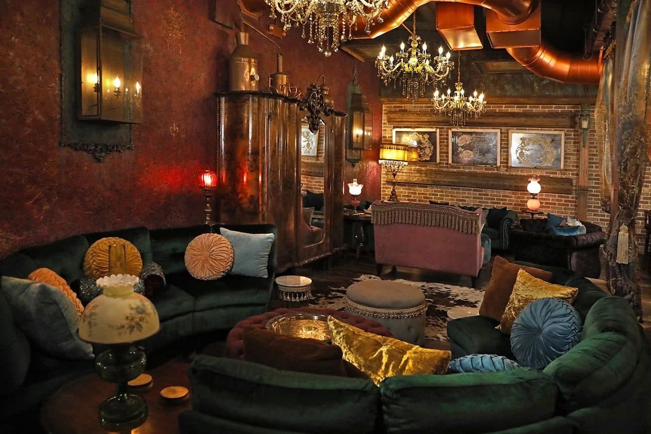 Interior of a moody speakeasy-style lounge (CC Social Club): green velvet sectional, gold/teal cushions, chandeliers, brick walls, red and amber lamp light; wide-angle architectural/hospitality shot.