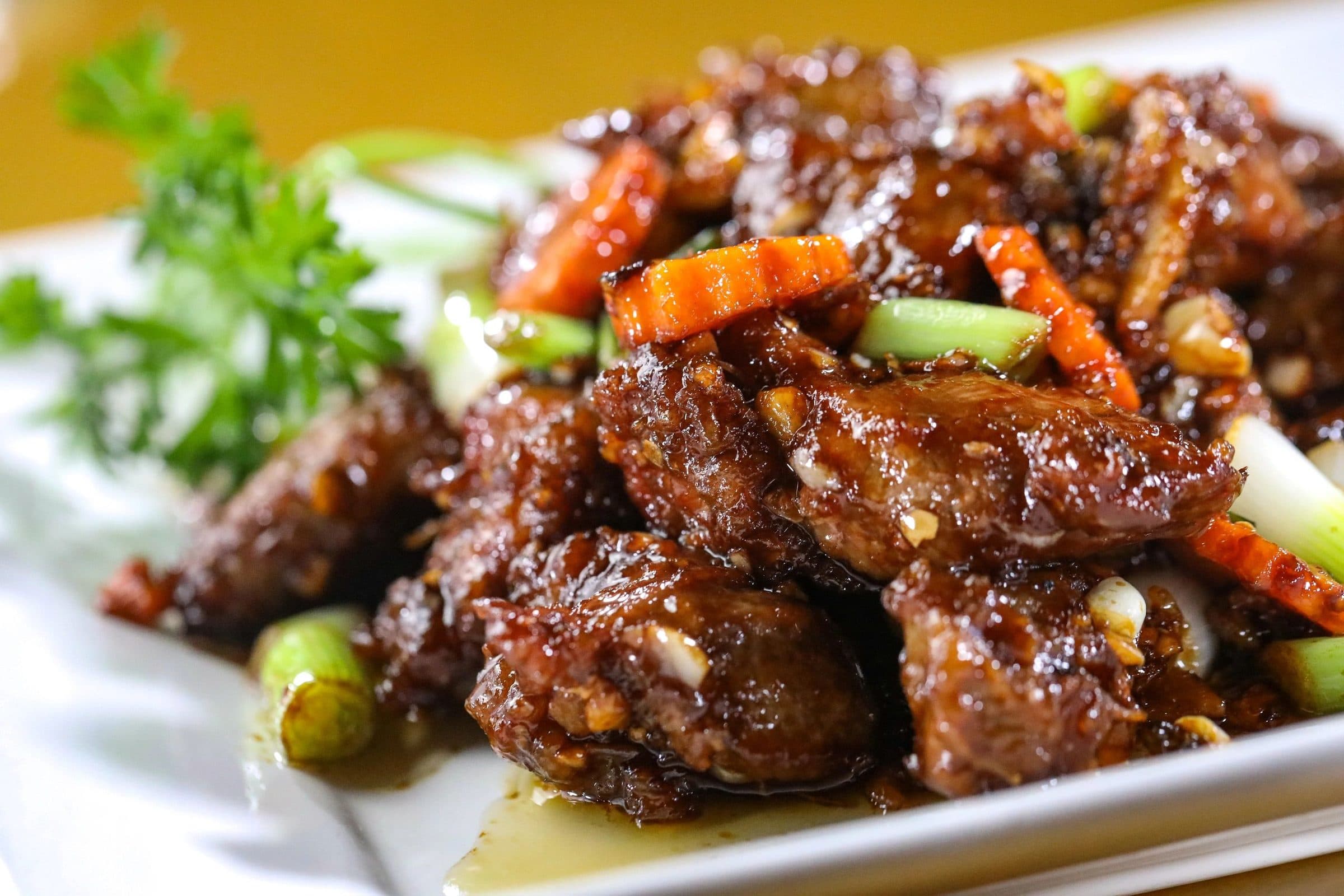 Close crop of sticky, deeply glazed fried chicken pieces (wings/nuggets) with roasted carrot batons, scallion segments and parsley on a white plate; warm yellow-orange background blur, glossy specular highlights, shallow depth of field.