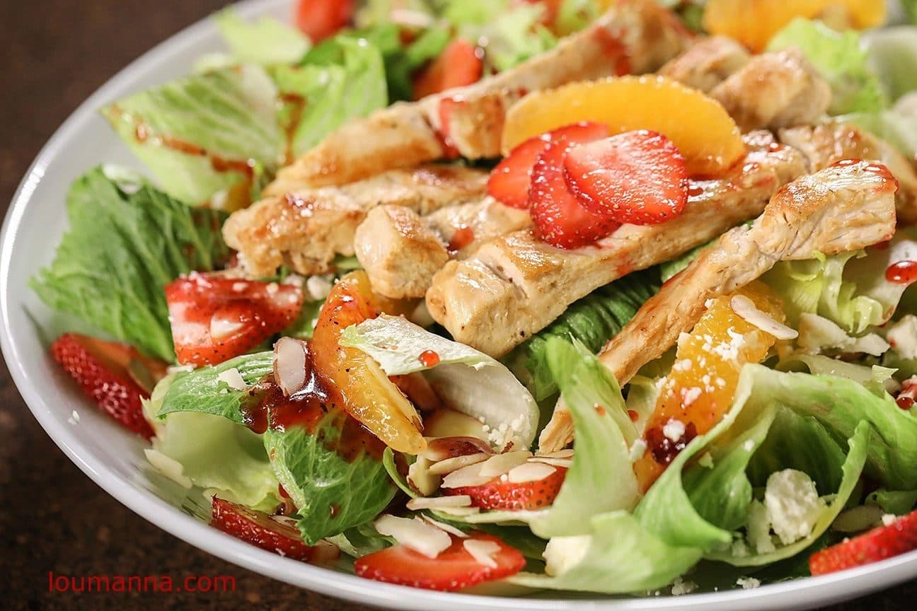 Overhead-angled grilled chicken salad in a white bowl: sliced chicken, strawberries, mandarin segments, almond slivers and romaine on a dark brown surface; bright even light; burned-in "loumanna.com" watermark lower left. Watermark would need cropping/retouch for hero use.