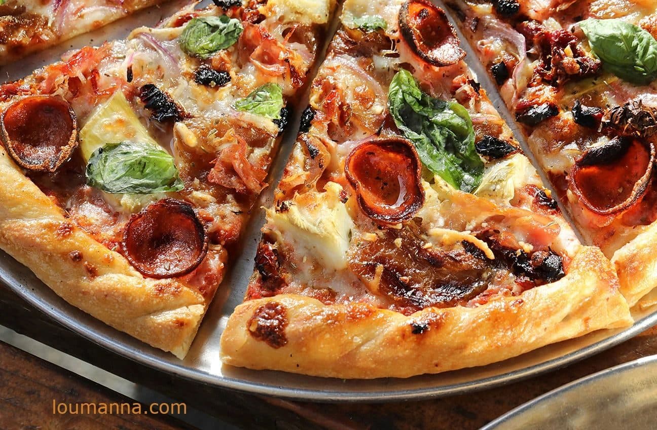 Tight overhead-angle crop of a sliced pepperoni pizza with fresh basil, artichoke and red onion on a metal tray (Noble Pie Parlor); warm light; burned-in orange "loumanna.com" watermark lower right. Watermark would need cropping/retouch for hero use.