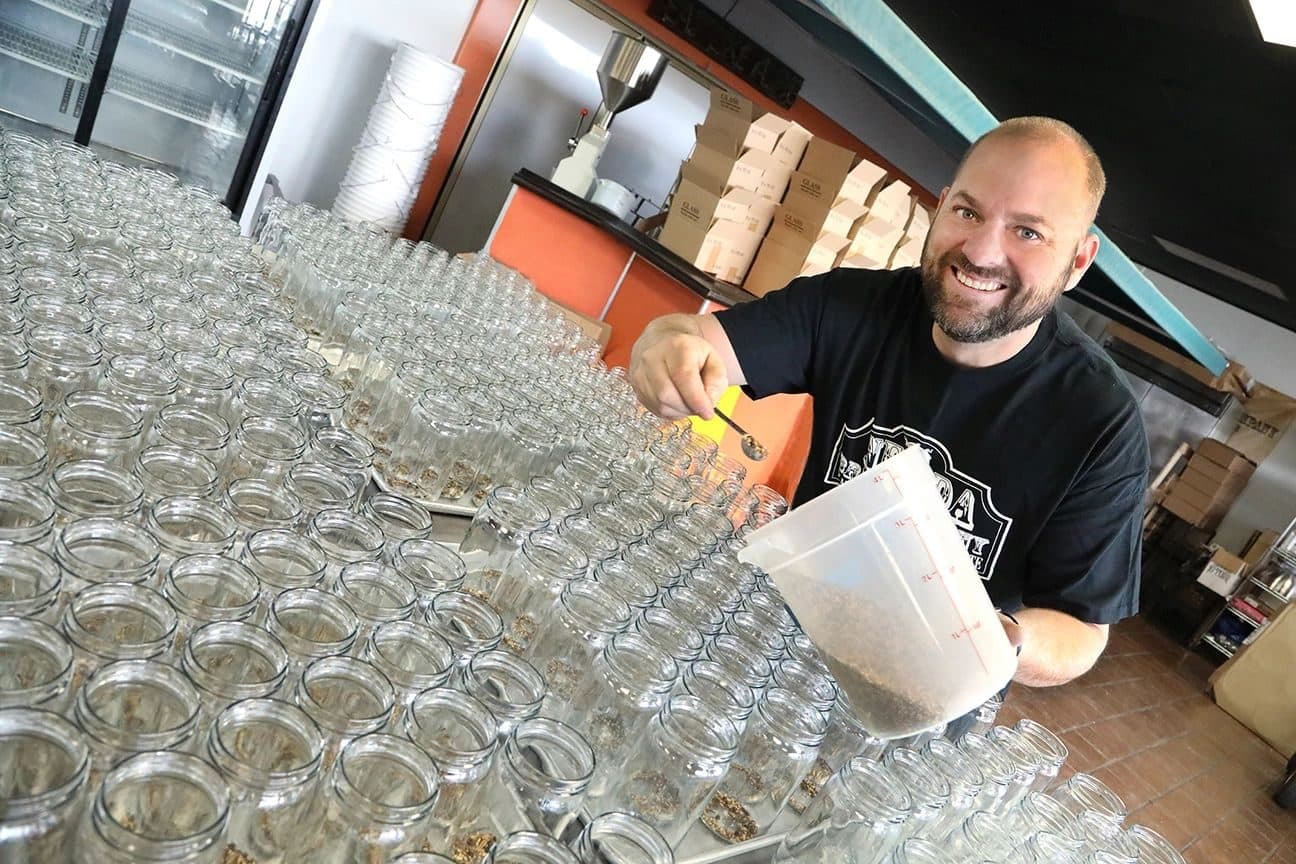 Environmental business portrait: bearded owner (Mat Soter per testimonial) smiling while spooning brine spices into rows of empty glass jars in a commercial kitchen; wide-angle, tilted frame, orange walls and boxed glassware.